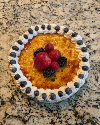 Crème brûlée cheesecake with a torched caramelized top, whipped cream and blueberry border, and fresh berries at the center