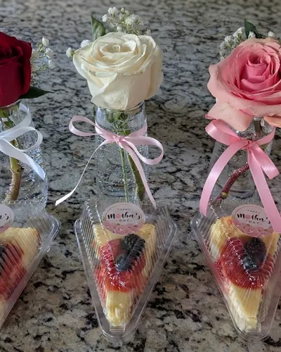 Three individual cheesecake slices topped with fresh berries, each paired with a single rose in a ribboned bud vase for Mother's Day