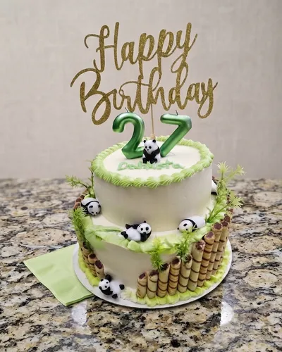 Two-tier birthday cake with a panda figurine on top, green number-27 candles, gold "Happy Birthday" topper, and small panda figures tucked into the tiers