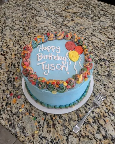 Round birthday cake with sky-blue buttercream, a piped rainbow-swirl border, and a personalized "Happy Birthday Tyson!" message
