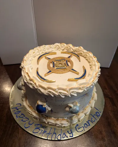 A single-tier birthday cake with a marbled blue, silver, and gold buttercream base, an ivory ruffled buttercream border with gold-dusted edges, a gold and blue emblem on top, and a "Happy Birthday Gancho!" message piped on the gold cake board