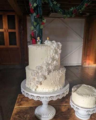 A two-tier rose-cascade wedding cake and a matching single-tier cake displayed on stands at a barn wedding venue, with an ivy and rose garland overhead